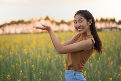 Optical illusion of young woman holding sun while standing on oilseed rape field against sky during sunset