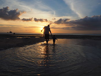 Rear view full length of father with child on shore against sky during sunset at beach