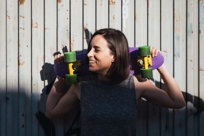 Smiling young woman holding skateboard while standing against wall