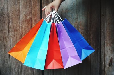 Cropped hand of person holding colorful shopping bags against wooden wall
