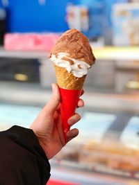 Soft cream with cocoa powder on top in waffle cone hold in a hand with soft focus of ice cream shop