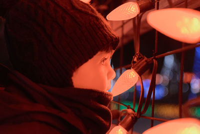 Close-up of boy looking through fence with illuminated lights at night