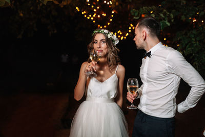 Portrait of couple holding wineglasses while standing outdoors at night