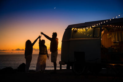 Friends toasting wineglasses while standing by camper van at beach against sky during sunset