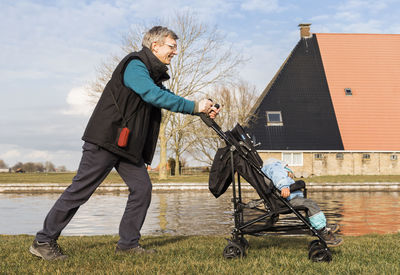 Side view of grandfather pushing baby stroller on field by lake