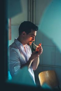 Side view of young man smoking cigarette at home seen through window