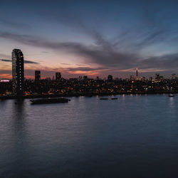 Illuminated buildings by river against sky at sunset