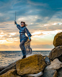 Rear view of man standing on rock at beach against sky during sunset