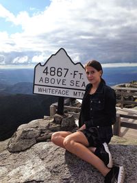 Portrait of woman sitting on rock by information sign against cloudy sky