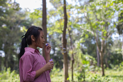 Cute girl blowing bubbles while standing against trees
