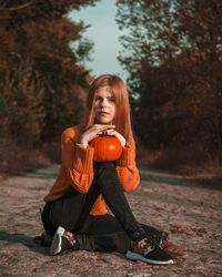 Full length of young woman holding pumpkin sitting against trees