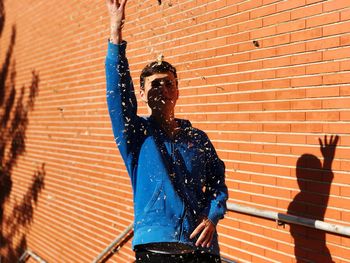 Young man throwing dry leaves while standing against brick wall