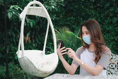 Young woman wearing mask using phone sitting outdoors