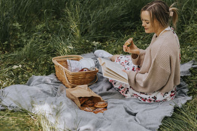 Smiling woman sitting on blanket and eating cinnamon bun while reading book