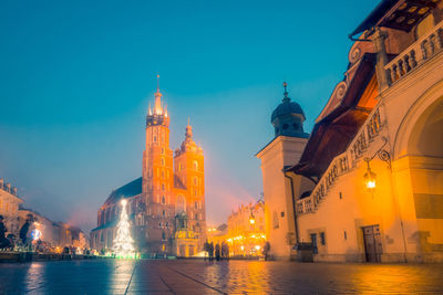 Spacious famous square with medieval buildings and church on paved path near glowing christmas tree on winter evening in poland