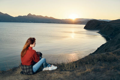 Woman sitting on shore against sky during sunset