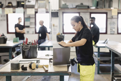 Side view of female student working on sheet metal with coworkers in background