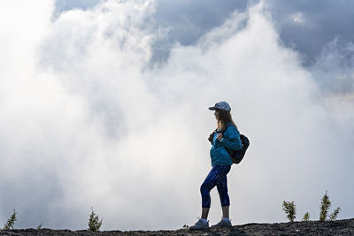 Young woman in cap blue sporty clothes with backpack in mountain against  white clouds, hiking 