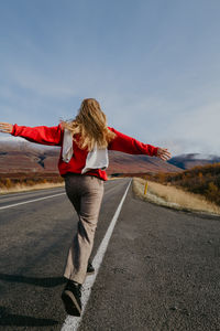 Woman in a red jumper and a scarf running on the road in iceland with arms spread