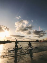 Rear view of friends paddleboarding on sea against sky during sunset