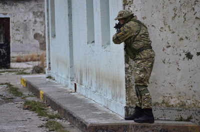 Army soldier aiming rifle while standing behind house in city