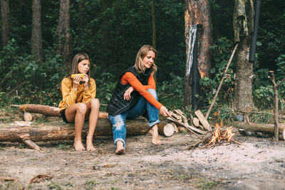 A mother with her teenage daughter is sitting on a tree near a campfire in the forest 