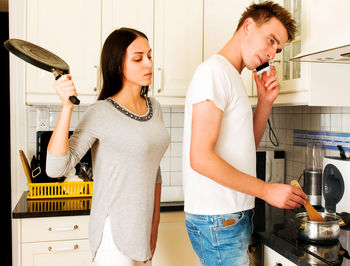 Young woman hitting man talking on smart phone in kitchen