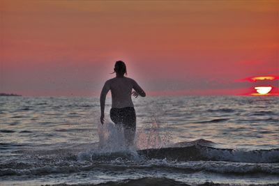Rear view of shirtless teenage boy running in sea at sunset
