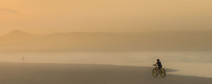 Boy riding bicycle on shore at beach against sky during sunset