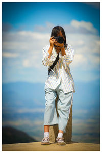 Low angle view of girl photographing while standing on rock against sky