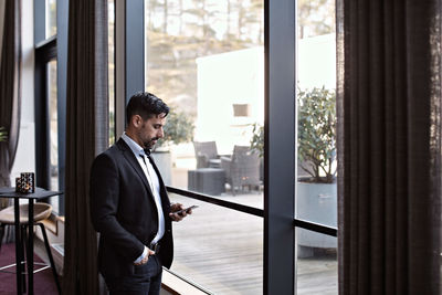 Businessman using phone while standing by window in office