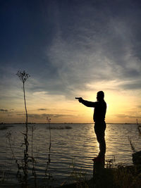 A silhouette of a man standing in sunset in front of a lake aiming a gun