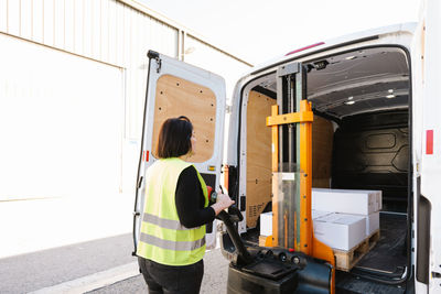 Side view of unrecognizable warehouse female worker in green uniform high vis vest loading boxes into opened truck with manual stacker on street