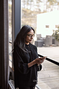 Businesswoman using smart phone while standing by window in office