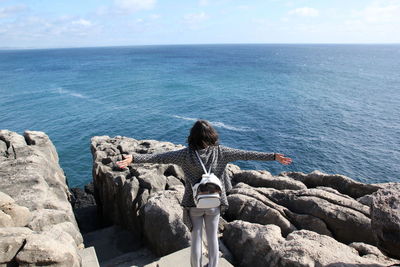 Rear view of woman standing with arms outstretched on rock at sea shore against sky