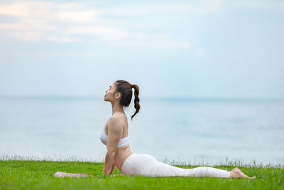 Side view of woman exercising on grass against sea