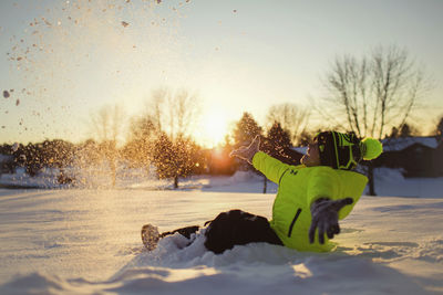 Boy playing with snow while sitting on field against sky during sunset