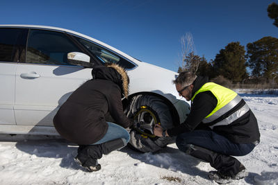 Male mechanic helping female driver with changing wheel of modern automobile on snowy road in winter