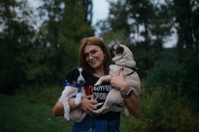 Portrait of smiling woman holding dog standing against trees