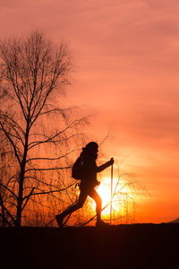 Silhouette woman walking by bare tree against orange sky