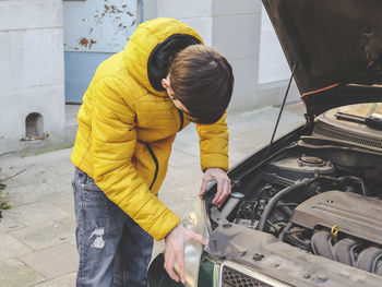 A young caucasian guy in a yellow jacket inspects an internal breakdown with an open car hood