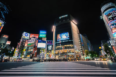 Illuminated city street by buildings against sky at night