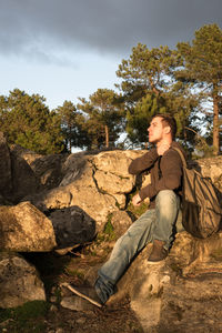 Young man looking away while sitting on rock against trees