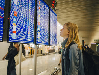 Side view of girl looking at departure board while standing at airport