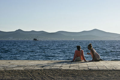 Rear view of couple sitting on retaining wall by sea against clear sky