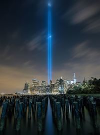 Wooden post in east river against light beams symbolizing world trade center in city against sky at night