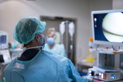 Back view of man in surgeon uniform looking at monitor while performing surgery on blurred background of modern operating theater