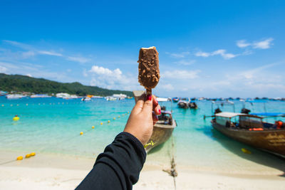 Cropped hand holding ice cream at beach against sky