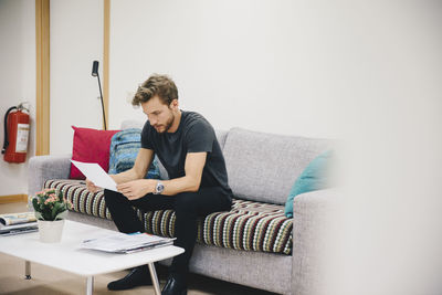 Young male patient reading document while sitting on sofa at hospital