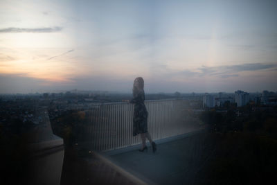 Woman standing by cityscape against sky during sunset